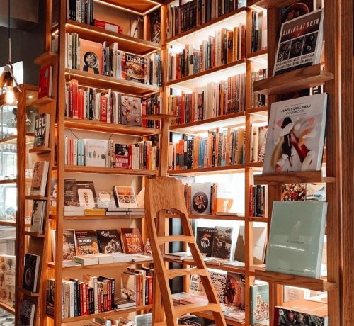 Estantería de madera rústica repleta de libros de cocina y gastronomía, con una escalera de madera integrada en el centro.